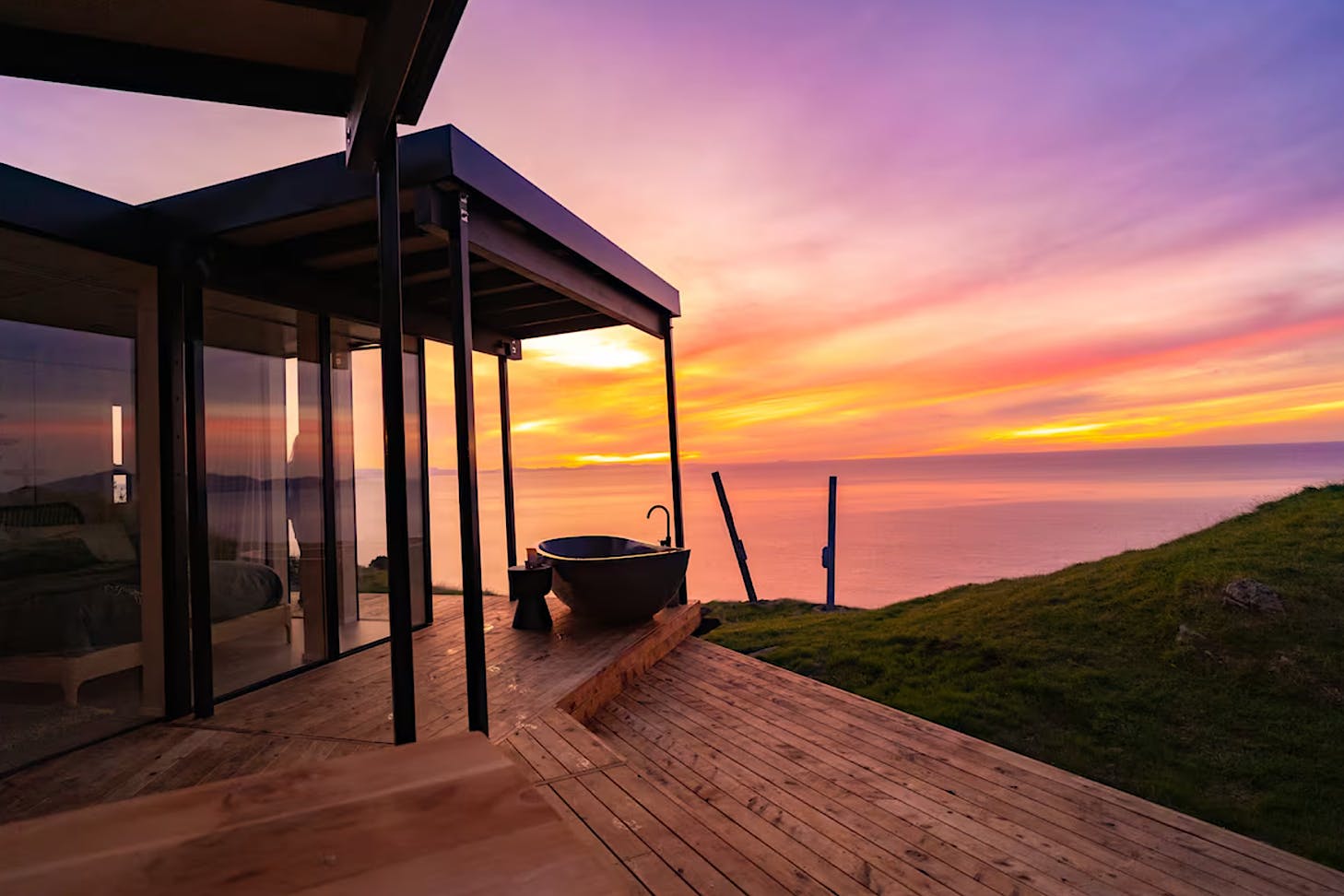 A vibrant purple and orange coastal sunset behind a modern Airbnb with an outdoor deck area and freestanding outdoor bath.